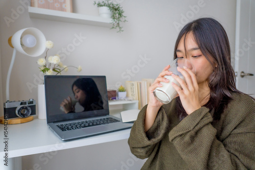 Young attractive Asian girl student drinking milk while sitting at study desk. Happy child enjoying a healthy drink during homework or online learning time in cozy home environment.