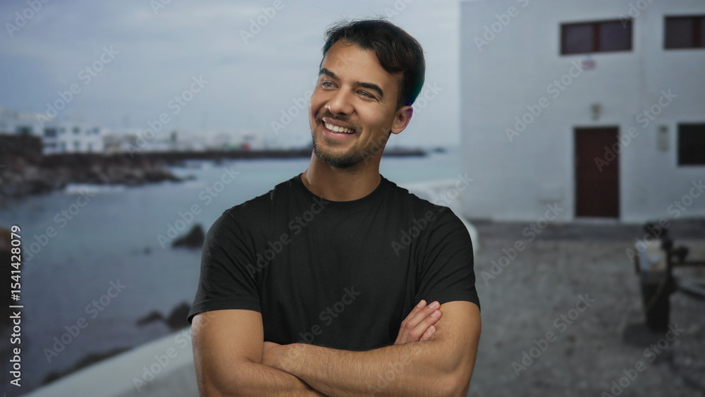Obraz premium Young hispanic man smiling confidently on seaside promenade with arms crossed, serene ocean backdrop enhancing outdoor setting.