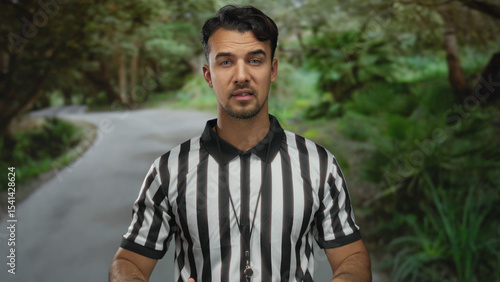Young hispanic man referee gesturing in park outdoor setting with green surroundings and blurred trees creating a natural atmosphere