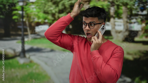 Young man in a red sweater speaks on a smartphone outdoors, surrounded by green park scenery.