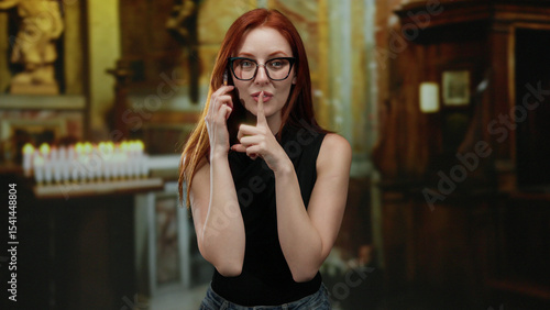 Young redhead woman gestures silence in a church setting, conveying discretion and calmness with lit candles in the background enhancing the serene indoor atmosphere.