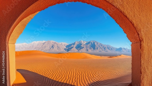 Desert vista through an archway.  A sandstone arch frames a panoramic view of a vast sand dune landscape stretching towards snow-capped mountains.  Sunlight bathes the dunes and mountains