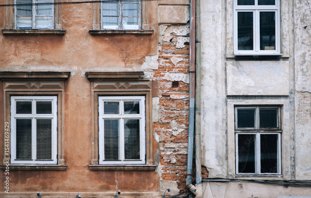 An old building with windows and a drainpipe is in a state of decay, revealing red bricks from under falling plaster. Concept of decline and aging. Lviv, Ukraine. An old building with windows and a drainpipe is in a state of decay, revealing red bricks from under falling plaster. Concept of decline and aging. Lviv, Ukraine.