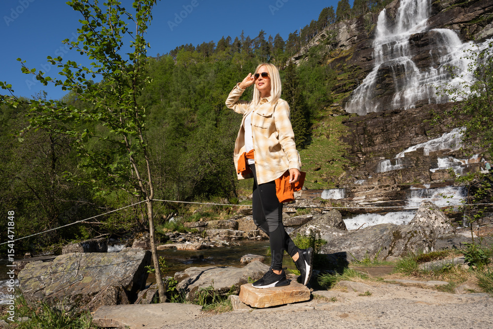 Naklejka premium A young female traveler is enjoying the beautiful sunny weather and a stunning waterfall view during a hiking trip in the mountains.