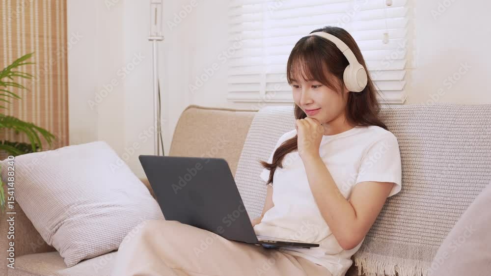 Asian woman smiling and working remotely with laptop and headphones on cozy sofa Asian woman smiling and working remotely with laptop and headphones on cozy sofa