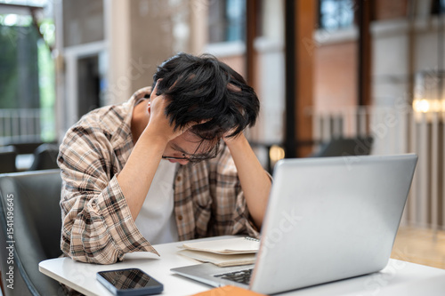 Man in plaid shirt holding hair thinking or stressing in front of laptop and notebook at cafe table.