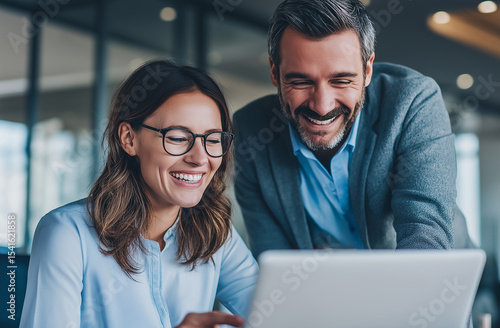 Two smiling professional business man and woman, sitting together at a desk in a modern industrial-style office, collaborating and looking at a laptop screen