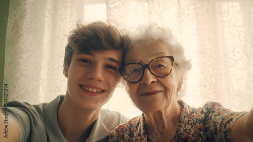 A teenager takes a cheerful selfie with his great-grandmother, both smiling widely with their heads touching. The elderly woman wears vintage glasses and a floral blouse, while the boy flashes a