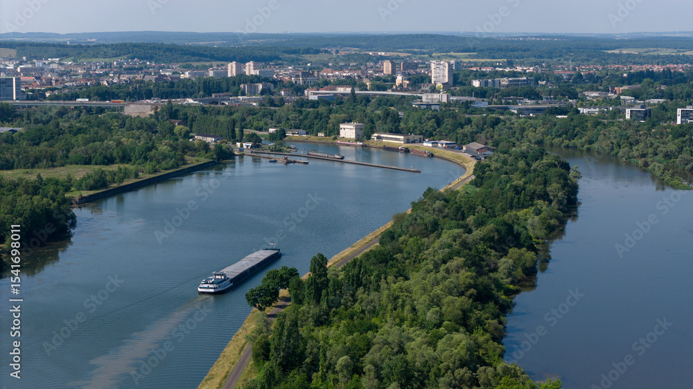 Fototapeta premium vue aérienne au dessus de la Moselle, une péniche avance sur l'eau. AU fond on aperçoit la ville de Thionville