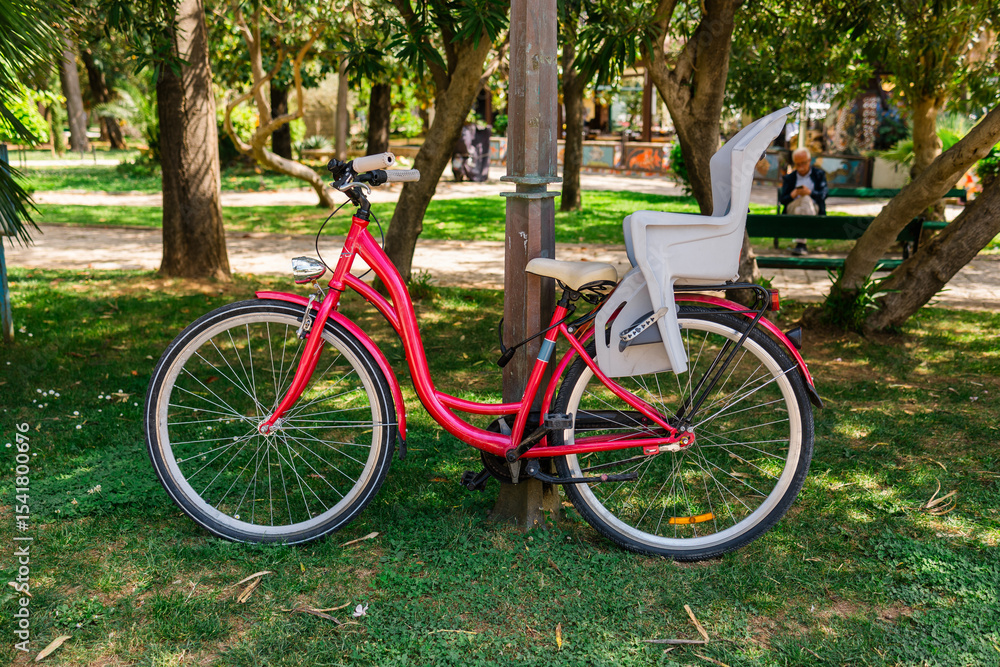Fototapeta premium Parked red bicycle with child seat. Family lifestyle, eco-friendly transport, and urban leisure in green outdoor spaces.