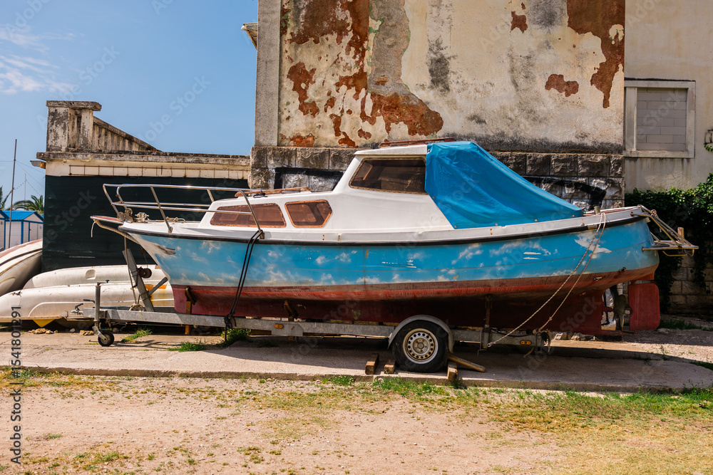 Fototapeta premium Old boat on trailer near weathered wall. Coastal lifestyle, fishing culture, and marine equipment storage