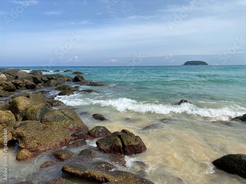 Rocky Tropical Beach with Clear Waves and Distant Island.Scenic view of a tropical beach with large rocks, gentle waves, and turquoise water under a clear sky.  Ideal for themes of travel, nature