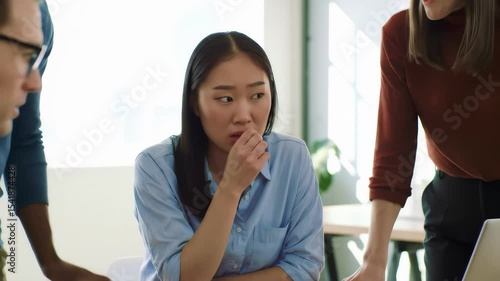 A nervous woman bites her nails during a tense team meeting, surrounded by concerned coworkers in a brightly lit office.
