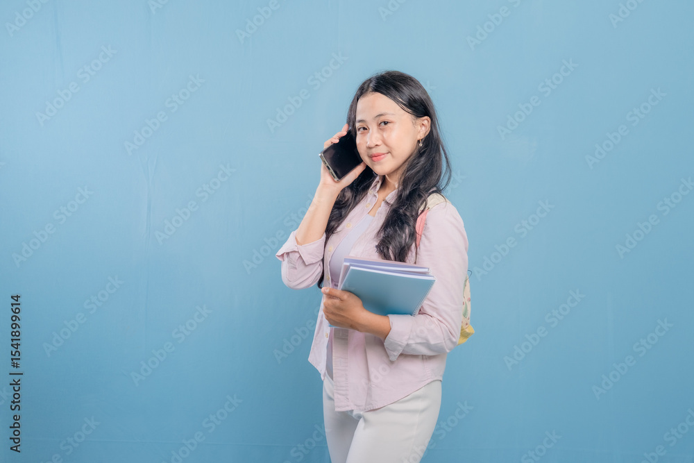 Fototapeta premium A woman is talking on her cell phone while holding a stack of books. She is smiling and she is in a good mood