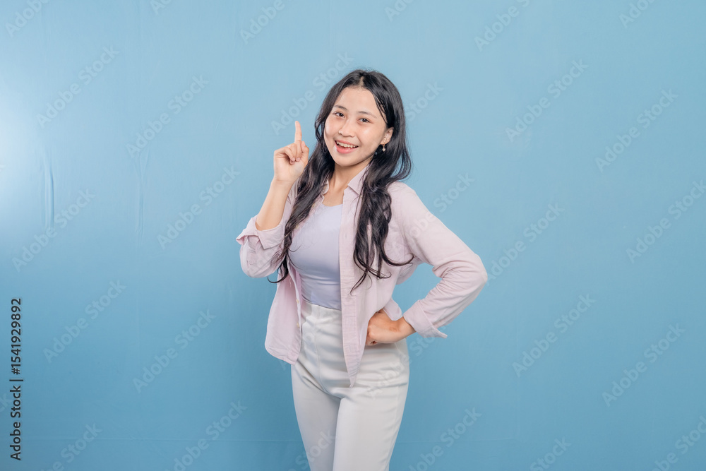 A woman is standing in front of a blue wall with a pink shirt and white pants. She is pointing her finger up and smiling A woman is standing in front of a blue wall with a pink shirt and white pants. She is pointing her finger up and smiling