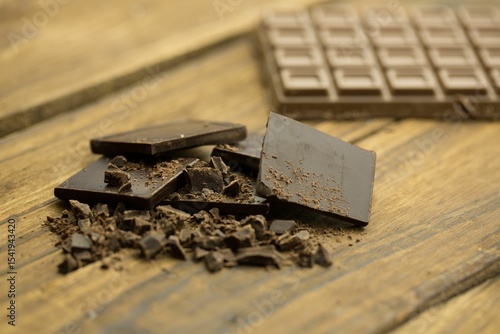 Dark chocolate pieces are resting on wooden tabletop in flat design, with blurred chocolate bar