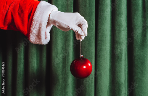 Santa's gloved hand holding a red Christmas ornament on green velvet background. Minimalist festive holiday concept.