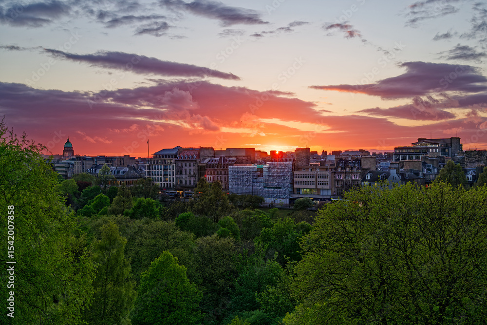 Fototapeta premium Beautiful sunset with orange pink sky above buildings on Princess street in Edinburgh, UK. Visible sun rays and colorful sky with clouds. Public park with green trees.