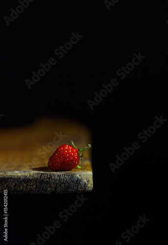 The image shows a single strawberry lying on the edge of a wooden surface. The background is black, which creates a contrast with the bright red berry illuminated by the light.