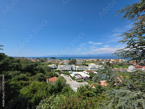 A scenic viewpoint overlooking a vibrant Greek tourist town, framed by green trees and the sea.