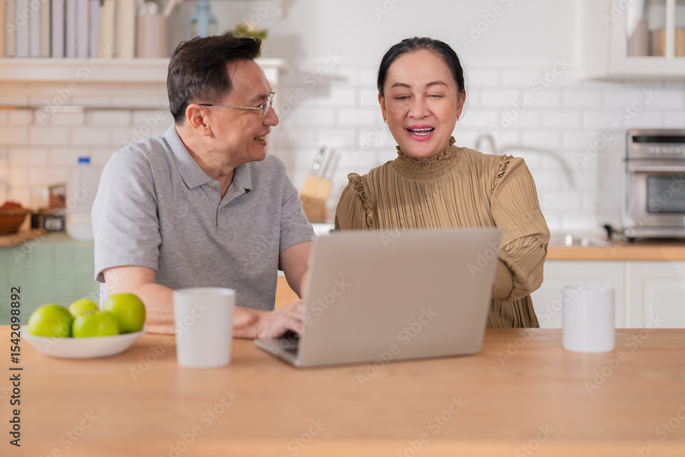 Asian senior couple sitting at the kitchen dining table, smiling warmly as they video call their grandson to celebrate his birthday online. Embracing technology, the grandparents use a digital tablet Asian senior couple sitting at the kitchen dining table, smiling warmly as they video call their grandson to celebrate his birthday online. Embracing technology, the grandparents use a digital tablet