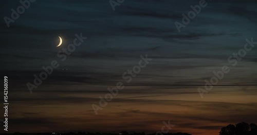 Timelapse of the nightfall scenery with dark orange and blue sky after sunset with wispy clouds and crescent moon slowly sinking toward the horizon