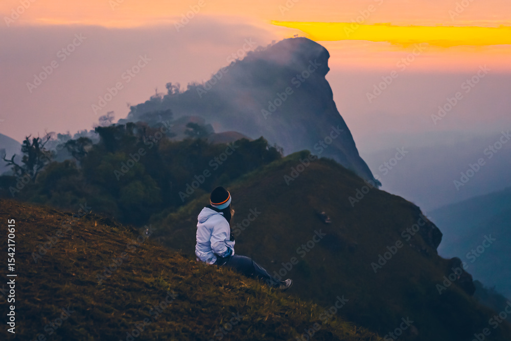 Fototapeta premium Photo of a woman sitting on a peak of the mountain during early sunrise. The sky is clear with warm tones of orange, yellow, and pink. Feeling fresh and free. The idea for trekking wallpaper.