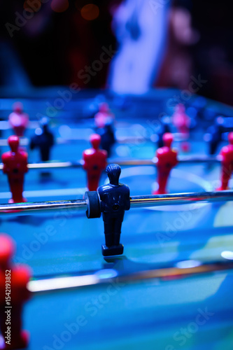 Table football setup in a bar atmosphere. The frame captures red and blue player figures on the game field, with a sharp focus on the front blue figure