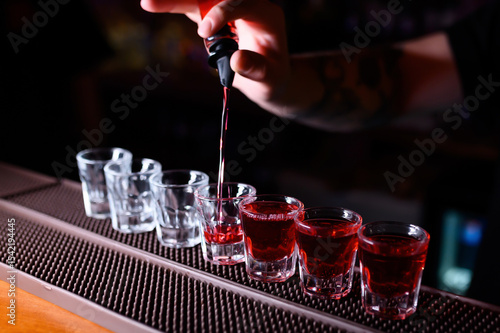A bartender is pouring bright red liqueur into a line of shot glasses on the bar counter. Half of the glasses are already filled, creating a vivid visual contrast between the empty and full ones
