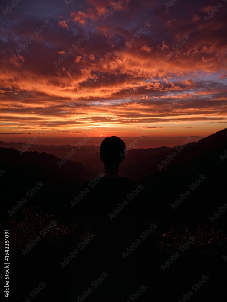 Silhouette of a person overlooking a vibrant sunset with orange and purple hues in the sky above mountains Silhouette of a person overlooking a vibrant sunset with orange and purple hues in the sky above mountains