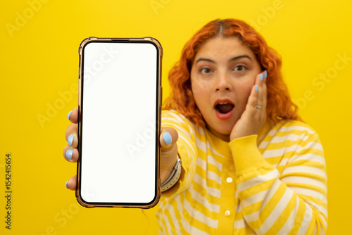 Young freckled woman with curly red hair with her hand on her face making a surprised expression showing her cell phone with a blank screen on a yellow background with copyspace