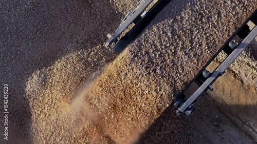 Top-down aerial shot following a mobile stone crusher's conveyor belt as it pours a steady stream of gravel, which is then moved and managed by a large wheel loader.
