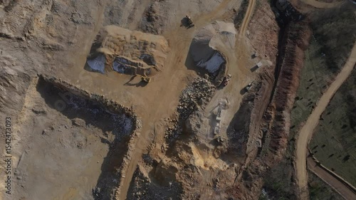 Top-down aerial view of a large open-pit quarry with heavy machinery. Excavators and loaders process stone, creating sand and gravel stockpiles for construction.
