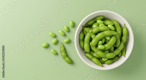 Top view of fresh green edamame beans in a white bowl with some scattered pods and soybeans on a pastel green background. Healthy plant-based protein snack or appetizer