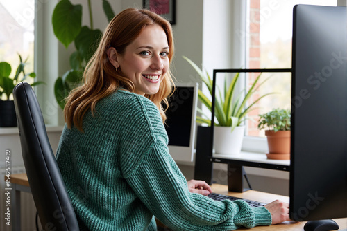 Woman looking over her shoulder and smiling while working at computer in bright, green office with many plants, enjoying positive and healthy work environment
