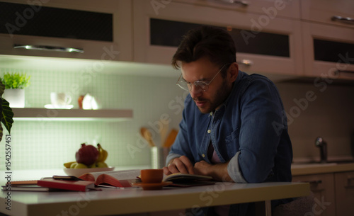 Obraz na płótnie A focused young man in glasses reads a book at a kitchen table at night, surrounded by open books, a cup of coffee, and warm ambient lighting, creating a cozy and studious atmosphere