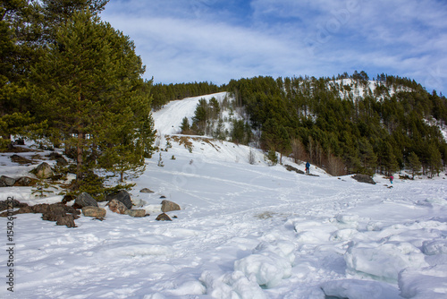  A frozen river or lake bordered by snowy hills and pine-covered slopes. Rocks dot the icy foreground under a partly cloudy sky, creating a serene winter mountain landscape.