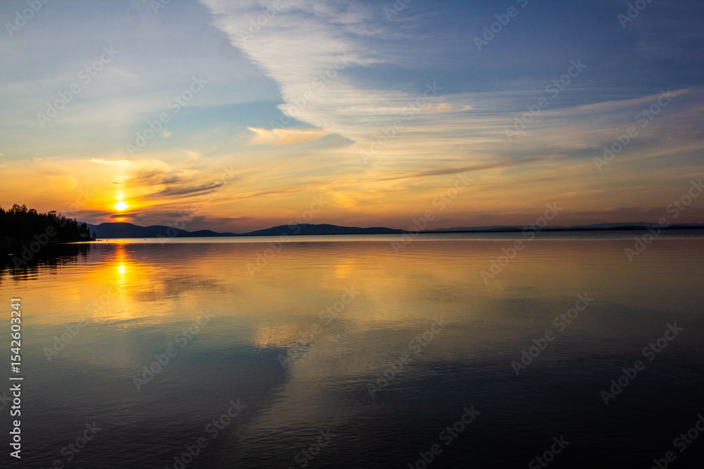 Obraz premium Stunning sunset over a calm lake with golden reflections, mountains on the horizon, and forest silhouette. Perfect for travel, nature backgrounds, peaceful landscapes, eco tourism, and inspiration.
