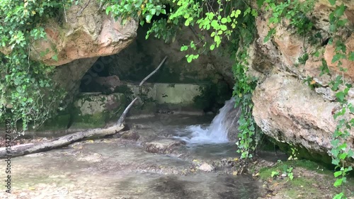 A small stream of water flows through a rocky cave. The water is clear and the rocks are brown. The scene is peaceful and serene