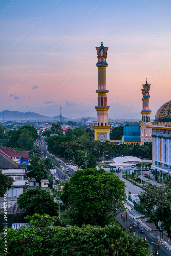 Obraz premium Golden twilight blankets the majestic mosque in Mataram city, Lombok, highlighting elegant minarets, vibrant streets, and surrounding hills under a serene pastel sky. Masjid Islamic Center Mataram.