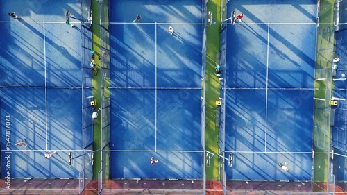 Aerial view of three vibrant blue padel courts, captured by a drone. Several players are actively engaged in padel matches, moving energetically across the courts. The scene is framed with strong