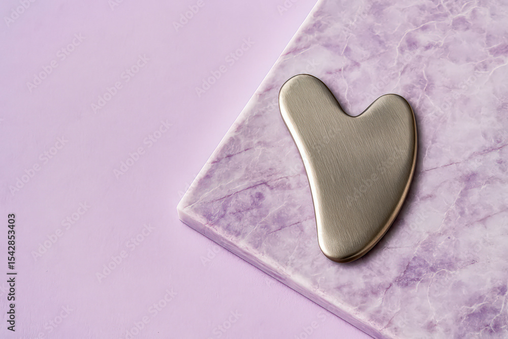 Fototapeta premium Elegant stainless steel gua sha facial massage tool displayed on a textured purple marble surface with a soft lavender backdrop, representing luxury skincare, beauty rituals, and holistic wellness.