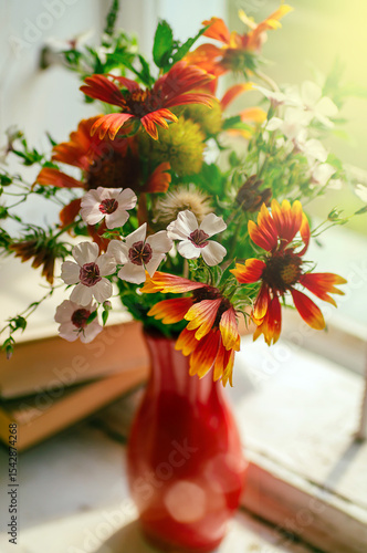 Bright bouquet of flowers on the window in a red vase in the rays of the sun