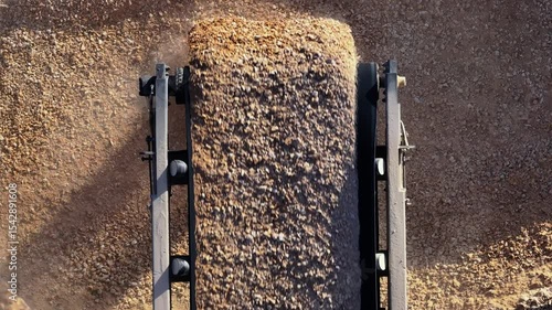 Top-down aerial view of a conveyor belt dropping crushed stone onto a large pile, with an excavator operating nearby at a quarry.
