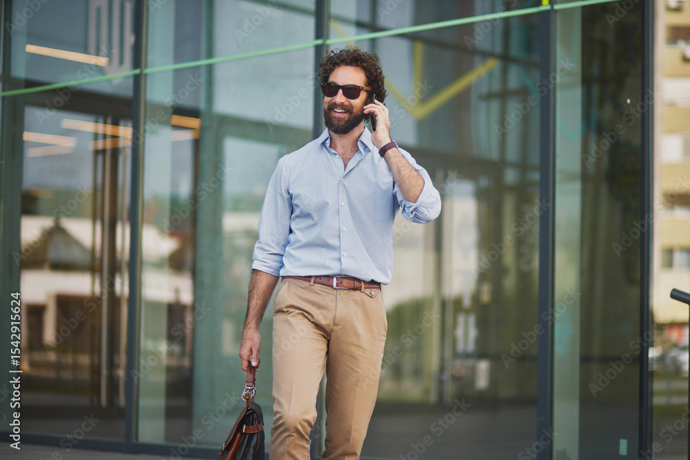 Fototapeta premium A man in business attire walks with a briefcase while engaged in a phone conversation outside a contemporary office building, showcasing a dynamic work environment.