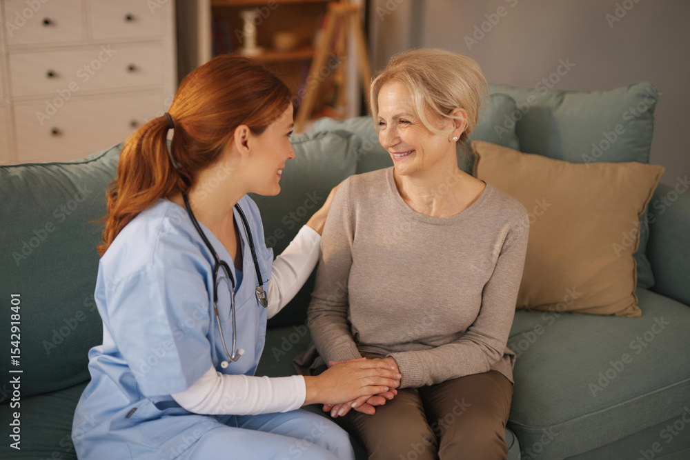 Obraz premium A redhead young nurse in blue scrubs comforts a blonde senior woman sitting on a teal couch inside a living room. The nurse holds the woman's hands, and both are smiling.