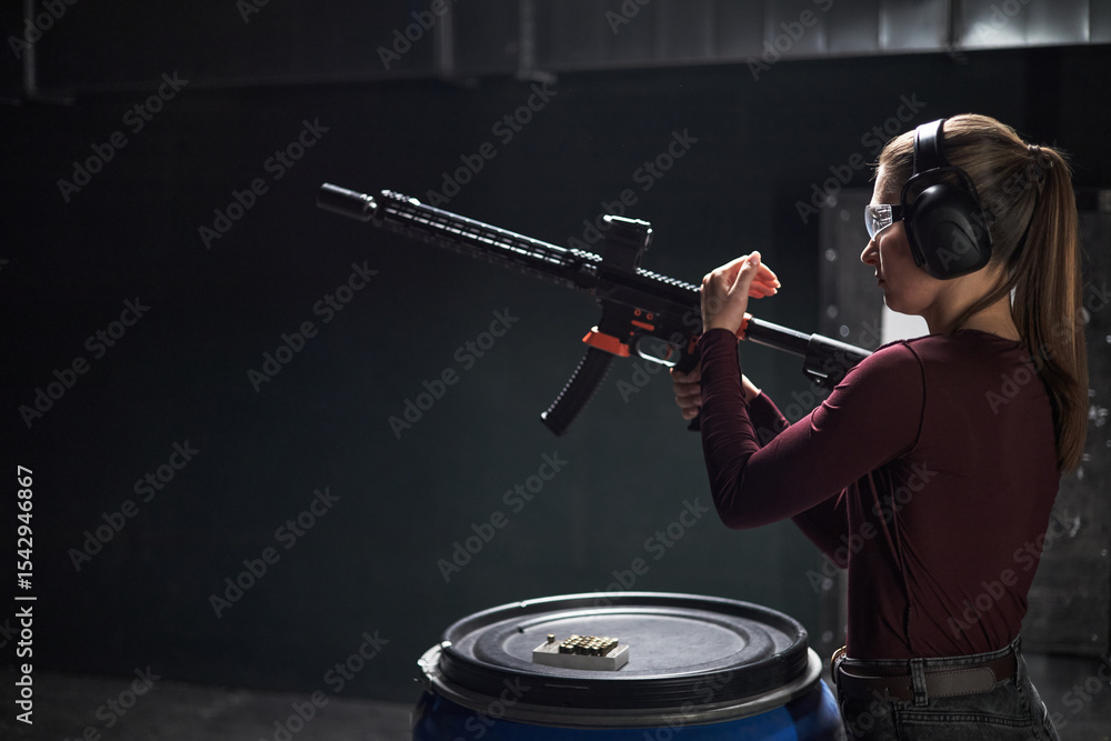 Fototapeta premium Young woman wearing protective goggles and earmuffs loading semi automatic rifle at indoor shooting club range, standing beside blue barrel with ammunition visible on top