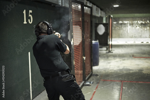Middle aged man shooting handgun at indoor range, wearing protective earmuffs, standing in firing position with visible smoke from weapon, practicing marksmanship