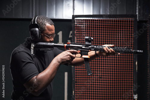 Tablou pe pânză Black middle aged man aiming rifle at indoor shooting range, wearing protective