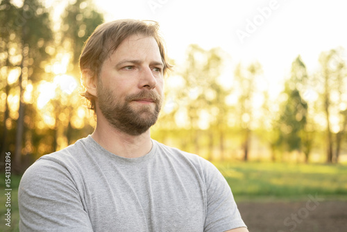 Portrait of handsome young man in gray t-shirt outdoors. Male with long hair and beard feel happy, calm and serious on Green nature sunset background. Man looking away into distance. Diversity People